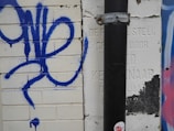 A section of a wall featuring blue graffiti on white bricks with a vertical black pipe partially obscuring a carved section in the wall. There is evidence of paint chipping and more graffiti in different colors on the right side.