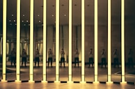 A modern art installation featuring tall mirrored panels with glowing edges. The reflections create multiple images of a person standing inside the room, capturing the interplay of light and shadow. The setting appears to be an exhibition space with a minimalist design.