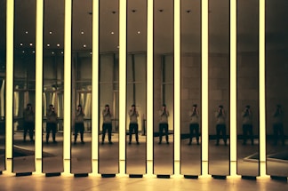 A modern art installation featuring tall mirrored panels with glowing edges. The reflections create multiple images of a person standing inside the room, capturing the interplay of light and shadow. The setting appears to be an exhibition space with a minimalist design.
