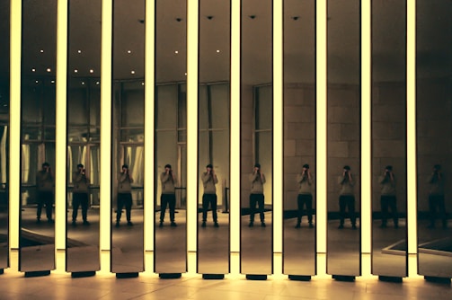 A modern art installation featuring tall mirrored panels with glowing edges. The reflections create multiple images of a person standing inside the room, capturing the interplay of light and shadow. The setting appears to be an exhibition space with a minimalist design.