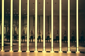 A modern art installation featuring tall mirrored panels with glowing edges. The reflections create multiple images of a person standing inside the room, capturing the interplay of light and shadow. The setting appears to be an exhibition space with a minimalist design.