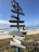 A colorful signpost stands on a beach, with several directional arrows pointing to various international locations. The sea and sky form a serene backdrop, with gentle waves rolling onto the shore.