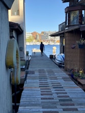 A person walks a dog along a wooden dock bordered by houseboats on a sunny day. In the distance, there are buildings and a marina with several sailboats lined up by the water.