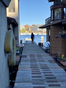 A person walks a dog along a wooden dock bordered by houseboats on a sunny day. In the distance, there are buildings and a marina with several sailboats lined up by the water.