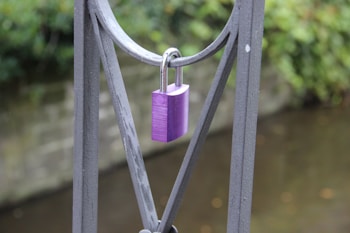 A purple padlock is attached to a metal fence with a river or water body in the blurred background. The metal fence has a geometric design and the scene conveys a sense of tranquility.