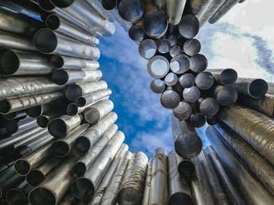 Numerous metal pipes arranged in an artistic formation with an upward view towards the sky. The pipes vary in size and create a dynamic, industrial structure set against a backdrop of blue sky with scattered clouds.