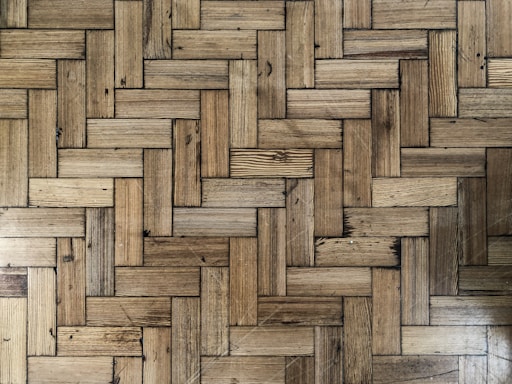 A close-up view of a wooden parquet floor with a herringbone pattern. The wood has various shades of brown and displays natural grain and knots, providing a rustic appearance.