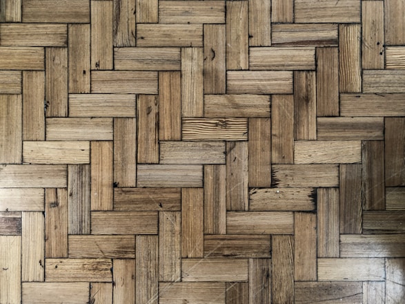 A close-up view of a wooden parquet floor with a herringbone pattern. The wood has various shades of brown and displays natural grain and knots, providing a rustic appearance.