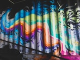 Vibrant graffiti art covers a corrugated metal surface, featuring swirling lines and bold colors. The design includes shades of blue, green, pink, orange, and yellow, blending into each other in an abstract manner. The graffiti displays energy and movement, with some drips and splashes suggesting dynamic creation.