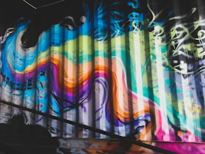 Vibrant graffiti art covers a corrugated metal surface, featuring swirling lines and bold colors. The design includes shades of blue, green, pink, orange, and yellow, blending into each other in an abstract manner. The graffiti displays energy and movement, with some drips and splashes suggesting dynamic creation.
