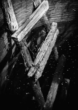 Old wooden beams partially submerged in water, with a textured surface and visible cracks. The reflection creates an abstract pattern, and the lighting emphasizes the grain and wear of the wood.