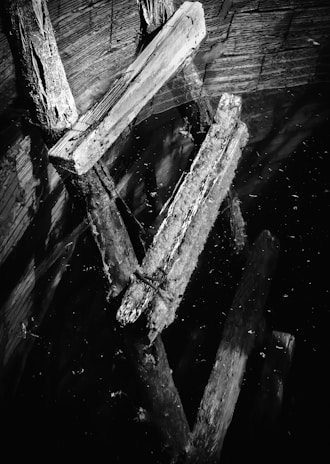 Old wooden beams partially submerged in water, with a textured surface and visible cracks. The reflection creates an abstract pattern, and the lighting emphasizes the grain and wear of the wood.