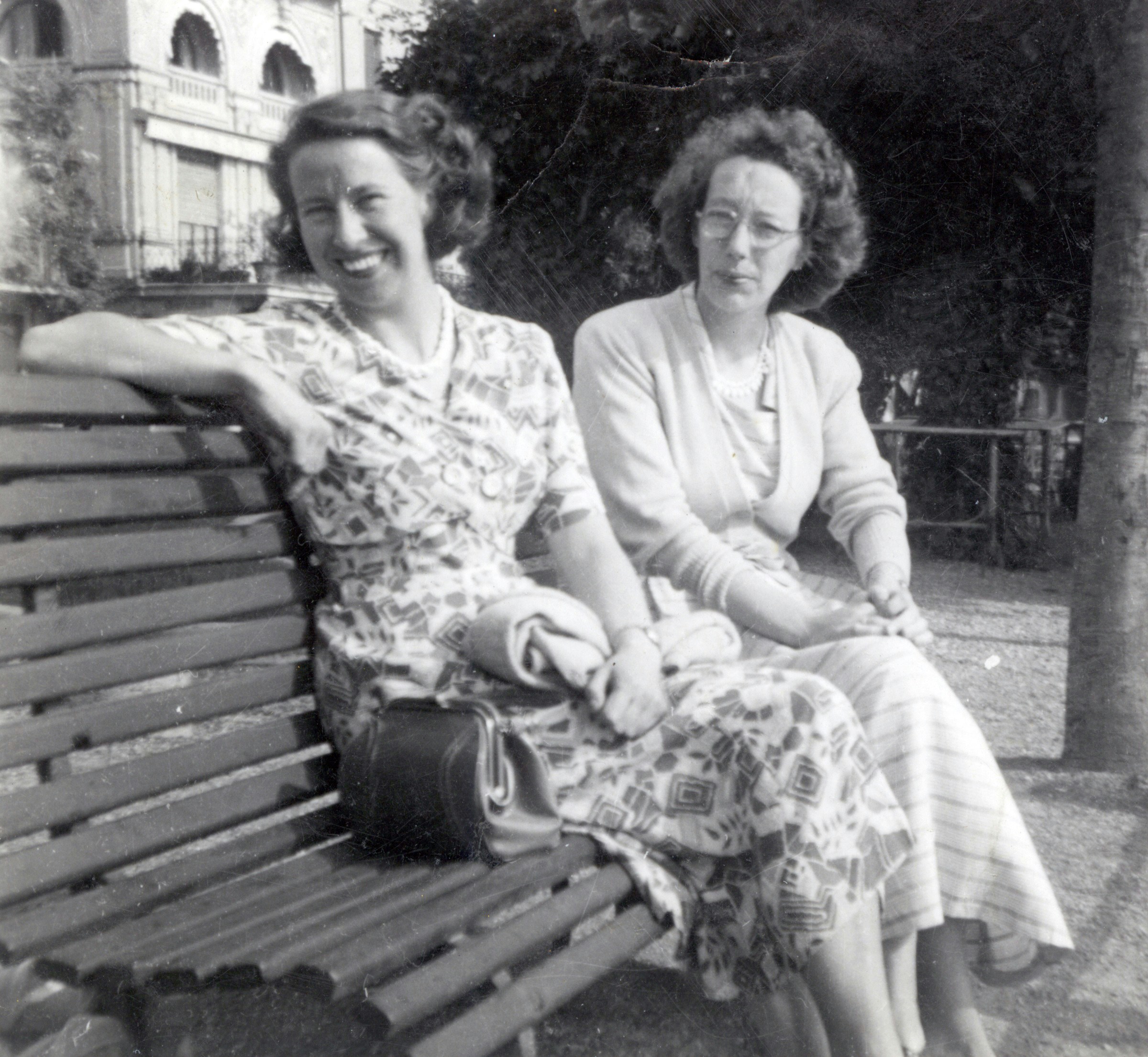 Two elderly women laughing together on a park bench, one adjusting the other's collar, warm afternoon light