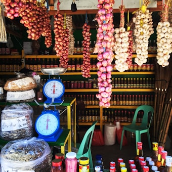 A market stall is filled with various goods including hanging bunches of garlic and onions. The shelves in the background display numerous jars and bottles of sauces and condiments. The stall also includes two blue mechanical weighing scales, plastic bags of dried goods, and a few green plastic chairs. The general atmosphere is colorful and bustling, with emphasis on the vibrant tones of the fresh produce and packaged items.