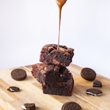 Two pieces of chocolate brownie stacked on a wooden surface, with a drizzle of caramel sauce pouring over the top piece. Surrounding the brownies are whole and broken sandwich cookies.