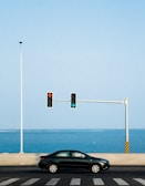 A black car is driving on a road with a view of the ocean in the background. A traffic light on a pole shows a green arrow pointing right and a red arrow pointing left. The sky is clear and blue, complemented by the ocean's hue.