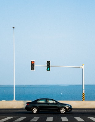 A black car is driving on a road with a view of the ocean in the background. A traffic light on a pole shows a green arrow pointing right and a red arrow pointing left. The sky is clear and blue, complemented by the ocean's hue.