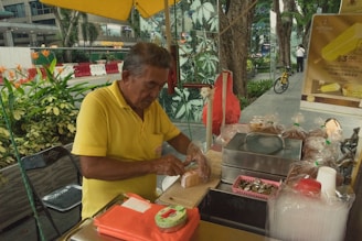 An elderly man, dressed in a yellow shirt, is preparing food at a street stall. The stall is equipped with various items like a bread roll, plastic wrap, a metal container, and a cash tray. In the background, there are lush green plants and trees lining the sidewalk. A bicycle and a poster advertising ice cream are also visible.