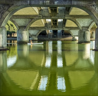 Under a large concrete bridge, a person navigates a white canoe across calm, greenish water. The arched architecture of the bridge is prominent, with reflections on the water's surface enhancing the scene's symmetry. Sunlight filters through openings, casting soft shadows.