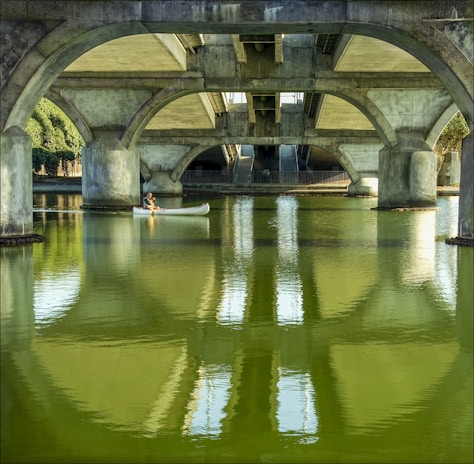 Under a large concrete bridge, a person navigates a white canoe across calm, greenish water. The arched architecture of the bridge is prominent, with reflections on the water's surface enhancing the scene's symmetry. Sunlight filters through openings, casting soft shadows.