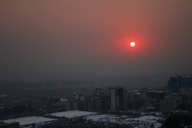 A city skyline is visible under a hazy, smog-filled sky. The sun, partially obscured by the pollution, glows a deep reddish-orange near the horizon. Tall buildings and urban infrastructure fill the foreground, with snow patches scattered in some areas, indicating a cold season.