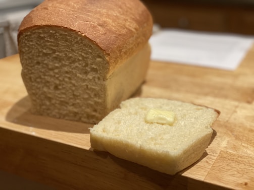 A freshly baked loaf of bread sits on a wooden cutting board. A slice has been cut and placed next to the loaf, topped with a small pat of butter.