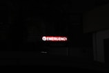 A bright red neon sign with the word 'Emergency' written, accompanied by a left-pointing arrow, stands out in a dark environment. Below, the word 'Parking' is faintly visible, indicating a parking area nearby.