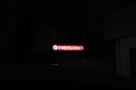 A bright red neon sign with the word 'Emergency' written, accompanied by a left-pointing arrow, stands out in a dark environment. Below, the word 'Parking' is faintly visible, indicating a parking area nearby.