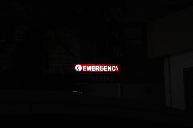 A bright red neon sign with the word 'Emergency' written, accompanied by a left-pointing arrow, stands out in a dark environment. Below, the word 'Parking' is faintly visible, indicating a parking area nearby.