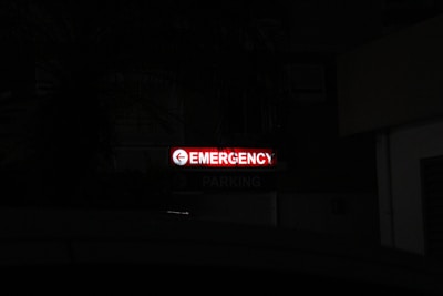 A bright red neon sign with the word 'Emergency' written, accompanied by a left-pointing arrow, stands out in a dark environment. Below, the word 'Parking' is faintly visible, indicating a parking area nearby.