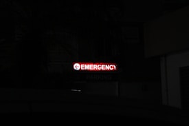 A bright red neon sign with the word 'Emergency' written, accompanied by a left-pointing arrow, stands out in a dark environment. Below, the word 'Parking' is faintly visible, indicating a parking area nearby.
