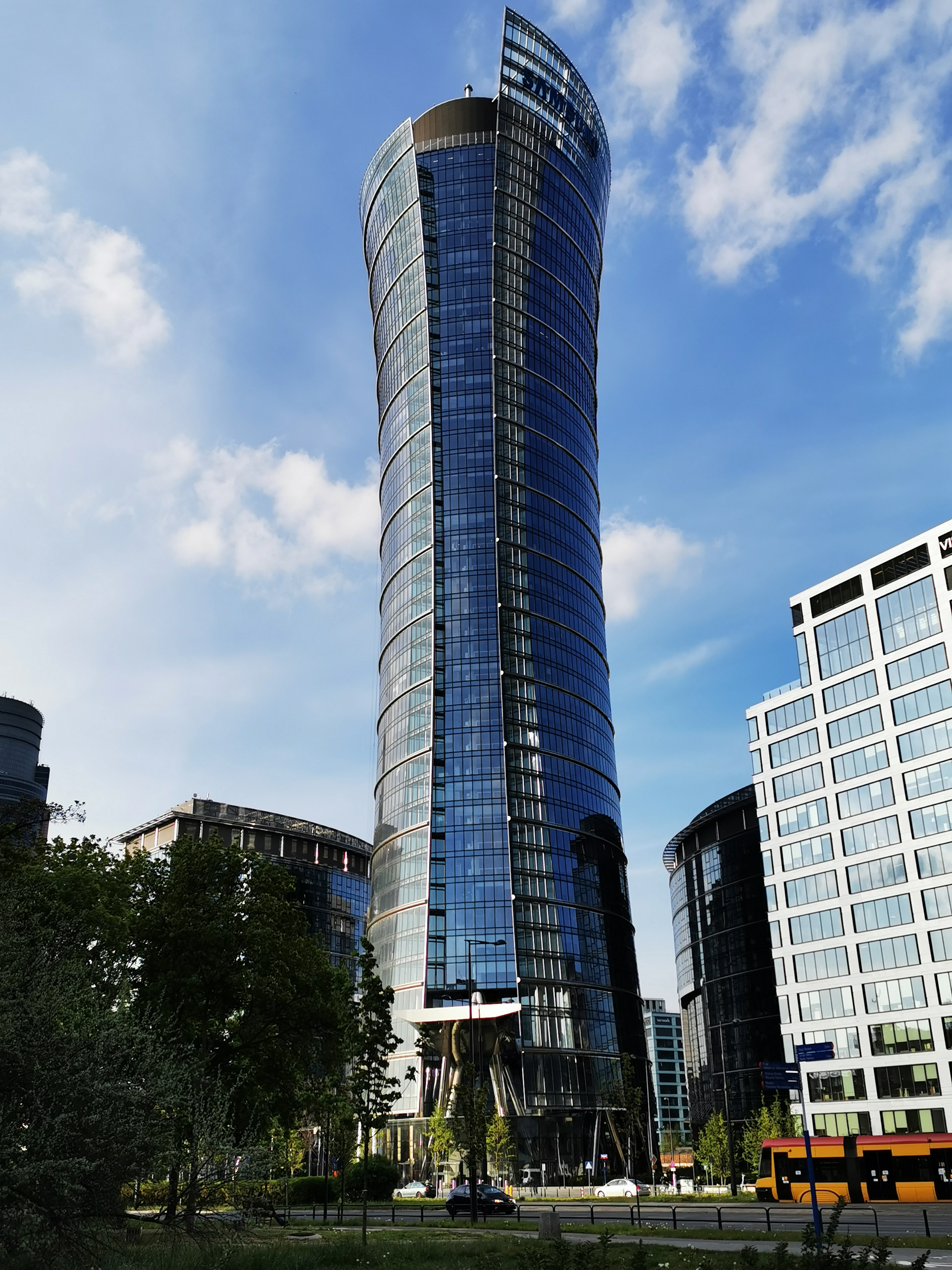 A curving glass skyscraper twists skyward above surrounding office towers under a blue sky