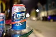 A can of Aroma beer is placed on a glass table with a city street in the background. The can features a blue and red design with the text 'Premium Quality' and 'Proudly Brewed Lager Beer'. The background is blurred, showing bokeh lights and a slightly visible street setting.
