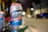 A can of Aroma beer is placed on a glass table with a city street in the background. The can features a blue and red design with the text 'Premium Quality' and 'Proudly Brewed Lager Beer'. The background is blurred, showing bokeh lights and a slightly visible street setting.