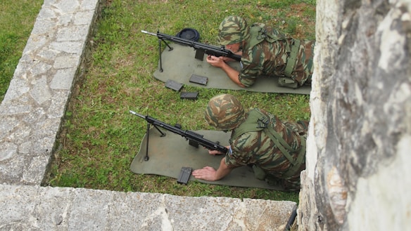 Two soldiers in camouflage uniforms are lying on mats with rifles in a grassy area surrounded by stone walls. They appear to be practicing or on guard duty.