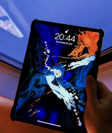 A hand holding a tablet displays a vibrant, abstract wallpaper with swirling patterns and contrasting colors, featuring blues, oranges, and whites. The tablet screen shows the time as 20:44 and the date as Donnerstag, 30. April. It is positioned near a window with a faint reflection visible.