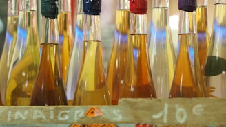 Several glass bottles filled with liquids of varying shades stand closely together. Each bottle is sealed with different colored tops, and they are arranged in a row within a wooden crate. Bright reflections and light create a colorful and vibrant display.
