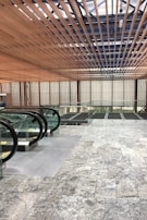 A modern architectural interior with a focus on geometric lines and natural materials. Wooden slats form a ceiling pattern, allowing light to filter through. Glass and metal railings accompany a series of escalators leading downward. The floor is covered in gray stone tiles, enhancing the contemporary feel.