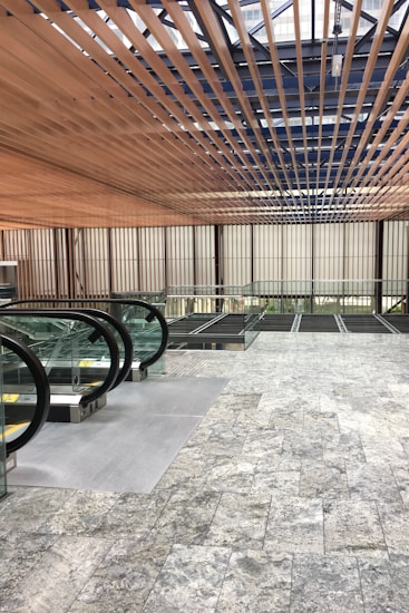 A modern architectural interior with a focus on geometric lines and natural materials. Wooden slats form a ceiling pattern, allowing light to filter through. Glass and metal railings accompany a series of escalators leading downward. The floor is covered in gray stone tiles, enhancing the contemporary feel.