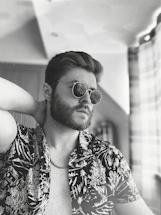 A person with sunglasses and a patterned shirt is leaning back with one hand behind the neck. The style is retro, with an emphasis on casual yet stylish attire. The black and white filter enhances the classic and timeless vibe of the scene.