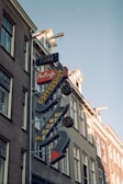 A vintage-style neon sign on the side of a building displays the words Internet Cafe, ProCafe, and Coffeeshop in bold letters. The sign features arrows and decorative elements, highlighting its retro aesthetic. The building itself is constructed with brick and has multiple windows. The sky is clear, indicating daytime.