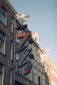 A vintage-style neon sign on the side of a building displays the words Internet Cafe, ProCafe, and Coffeeshop in bold letters. The sign features arrows and decorative elements, highlighting its retro aesthetic. The building itself is constructed with brick and has multiple windows. The sky is clear, indicating daytime.