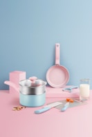 A pastel-themed kitchen setup featuring a set of utensils. There is a saucepan with a lid, a frying pan, a knife, a spatula, a peeler, and two measuring spoons. The background is a smooth gradient of pink and blue, with a glass of milk and some chopped vegetables adding a touch of realism.