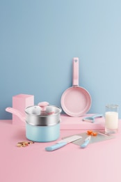 A pastel-themed kitchen setup featuring a set of utensils. There is a saucepan with a lid, a frying pan, a knife, a spatula, a peeler, and two measuring spoons. The background is a smooth gradient of pink and blue, with a glass of milk and some chopped vegetables adding a touch of realism.