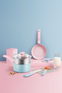 A pastel-themed kitchen setup featuring a set of utensils. There is a saucepan with a lid, a frying pan, a knife, a spatula, a peeler, and two measuring spoons. The background is a smooth gradient of pink and blue, with a glass of milk and some chopped vegetables adding a touch of realism.