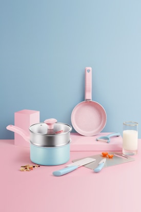 A pastel-themed kitchen setup featuring a set of utensils. There is a saucepan with a lid, a frying pan, a knife, a spatula, a peeler, and two measuring spoons. The background is a smooth gradient of pink and blue, with a glass of milk and some chopped vegetables adding a touch of realism.