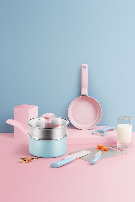 A pastel-themed kitchen setup featuring a set of utensils. There is a saucepan with a lid, a frying pan, a knife, a spatula, a peeler, and two measuring spoons. The background is a smooth gradient of pink and blue, with a glass of milk and some chopped vegetables adding a touch of realism.