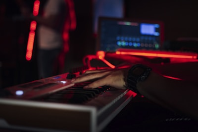 A person is playing a keyboard in a dimly lit room. The scene is illuminated by red LED lights, creating a moody atmosphere. A laptop displaying music software is visible in the background.