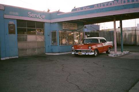 An old service station featuring a vintage red and white car parked under an awning. The building has a faded blue paint with signs indicating vehicle maintenance services, and the ground is paved with cracked asphalt.