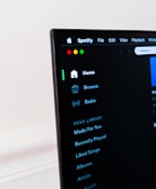 A computer screen displays the Spotify application, showing menu options such as Home, Browse, Radio, and Your Library, which includes Made For You, Recently Played, and Liked Songs. The application interface is sleek, with a dark background and white text, alongside green, yellow, and red window control buttons at the top left.